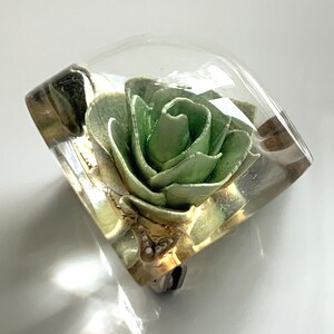 以下が含まれることがあります： 透明な正方形の樹脂製のブローチで、中に緑色の紙の花が入っています。花は、複数の層の花びらを持つ単一のバラです。