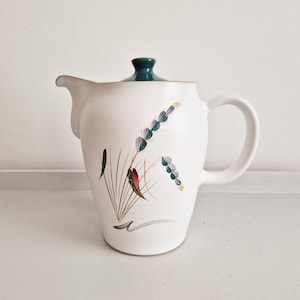 Peut inclure: Théière en céramique blanche avec un couvercle vert et un motif floral représentant des épis de blé bleus et verts et des feuilles rouges.