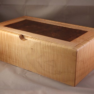 May include: A rectangular wooden box featuring a light-colored, wavy-grained wood body and a contrasting dark brown, smooth rectangular inlay on the top lid. The box has a small, semi-circular cutout on the front edge for opening the lid.