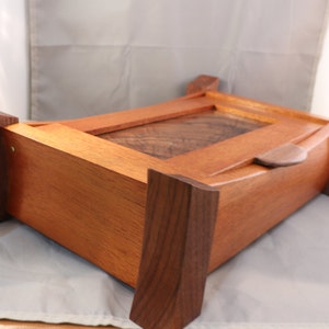 May include: A rectangular wooden box featuring a light reddish-brown body with a darker brown wood grain panel on the lid. The box is supported by four thick, dark brown wooden legs at the corners and includes a small, curved wooden handle on the lid.