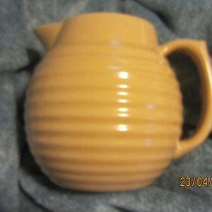 May include: A tan ceramic jug with a ribbed design. The jug has a handle and a spout.