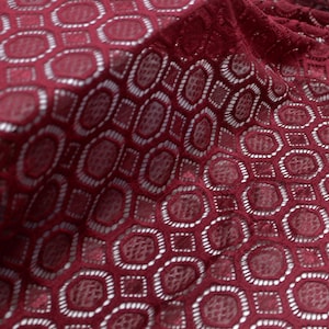 Könnte beinhalten: Nahaufnahme eines bordeauxfarbenen Spitzenstoffs mit einem sich wiederholenden geometrischen Muster. Das Design zeigt miteinander verbundene Achtecke und Quadrate, die eine offene, luftige Textur erzeugen. Die Farbe des Stoffes ist ein tiefes, sattes Rot.