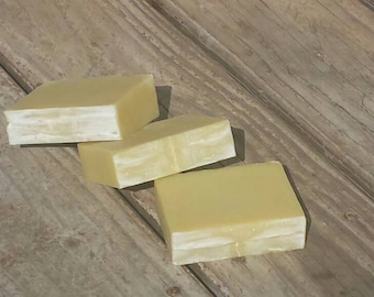 May include: Three bars of handmade soap, each with a light yellow color and a white layer on top. The soap bars are arranged on a wooden surface.