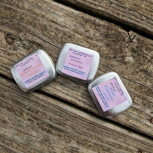May include: Three small, silver tins with pink labels. The labels have the text "granola girl" and the names of different lotion bars: "Optimistic Orange", "Lavender", and "Peppy Peppermint".
