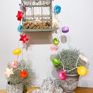 May include: A decorative display featuring a small, antique-style birdcage filled with corks, adorned with a string of colorful, illuminated lights. The lights include star and shell shapes in various colors, adding a whimsical touch.