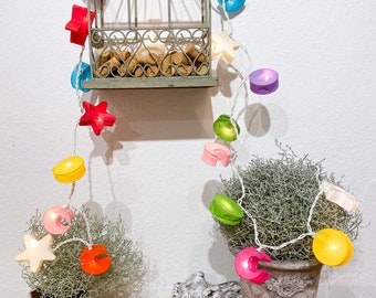 Puede incluir: Una exhibición decorativa con una pequeña jaula para pájaros de estilo antiguo llena de corchos, adornada con una cadena de luces de colores. Las luces incluyen formas de estrella y concha en varios colores, agregando un toque caprichoso.