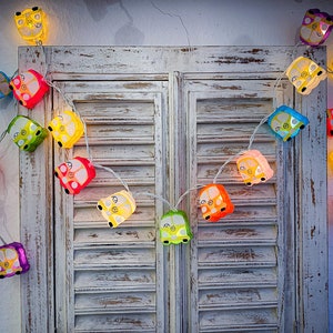 May include: A string of colorful lights shaped like vintage vans, hanging on a white wooden door with shutters. The lights are strung together with clear wire and each van has a different color.