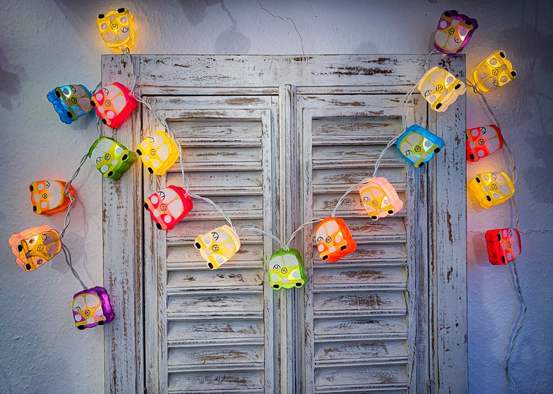May include: A string of colorful lights shaped like vintage vans, hanging on a white wooden door with shutters. The lights are strung together with clear wire and each van has a different color.