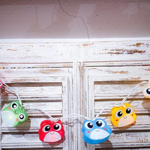 May include: A string of colorful frog-shaped lights with white wires. The lights are hanging on a white wooden shutter.