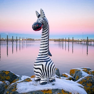Puede incluir: Una figurita de cebra en blanco y negro con una boca roja se encuentra sobre una roca cubierta de nieve y líquenes. El fondo es una puesta de sol rosa y azul sobre una masa de agua con postes de madera en el agua.