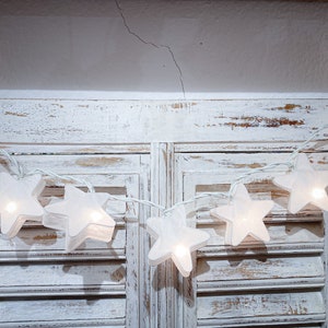 May include: A string of white star-shaped fairy lights. Each star is illuminated, creating a soft glow. The lights are strung on a white wire and are draped over a distressed white wooden surface, perfect for holiday or event decor.