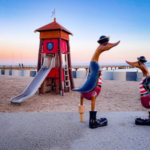 Könnte beinhalten: Holzenten-Skulpturen in Matrosenoutfits stehen auf einer Betonfläche in Strandnähe. Im Hintergrund befindet sich ein roter Rettungsturm mit Rutsche. Die Enten tragen schwarze Stiefel, Hüte und gestreifte Hemden. Der Himmel ist ein Farbverlauf aus Blau und Orange.