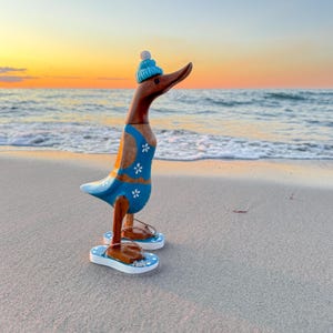 May include: A wooden duck figurine, painted with a blue swimsuit and a matching beanie. The duck is wearing brown flip-flops with white and blue soles. The background shows a beach at sunset.