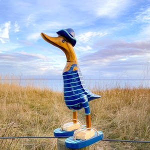 Könnte beinhalten: Holz-Entenfigur mit blau-weiß gestreiftem Hemd, blauem Hut und blauen Sandalen. Die Ente steht auf einem Holzpfosten mit einem Hintergrund aus trockenem Gras, einem Gewässer und einem bewölkten Himmel.