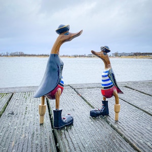 May include: Two wooden pirate duck figurines standing on a wooden dock. The ducks are wearing black and red pirate costumes with blue and white striped shirts. The larger duck is wearing a black hat with a gold trim. The smaller duck is wearing a black eye patch and a black hat with a gold trim.