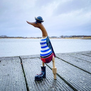 Puede incluir: Una figura de pato de madera vestida de marinero, con una camisa a rayas azules y blancas, pantalones rojos y un sombrero negro. El pato tiene una bota negra y está de pie en un muelle de madera cerca de una masa de agua.