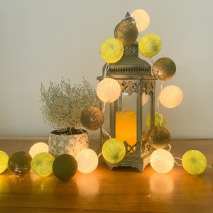 May include: A string of cotton ball lights with a mix of white, green, and brown balls. The lights are wrapped around a decorative metal lantern with a candle inside. The lantern is sitting on a wooden table with a small potted plant in front of it.