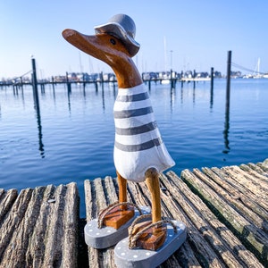 以下が含まれることがあります： グレーの帽子、グレーのストライプの白いシャツ、グレーのビーチサンダルを履いた木製のカモの置物。カモは、水面と背景にボートがある木のドックに立っています。カモのくちばしは茶色です。