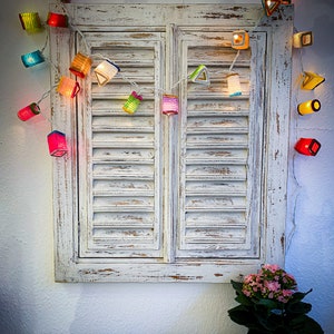 May include: A string of colorful, illuminated paper lanterns hangs above a distressed white wooden shuttered window. The lanterns are various shapes and colors, including red, yellow, blue, and green. A small potted plant with pink flowers sits below the window.