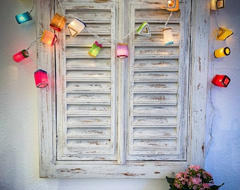 May include: A string of colorful, illuminated paper lanterns hangs above a distressed white wooden shuttered window. The lanterns are various shapes and colors, including red, yellow, blue, and green. A small potted plant with pink flowers sits below the window.