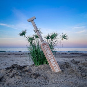 May include: A wooden sign with the word "HEIMATHAFEN" in white lettering, leaning against a patch of green grass on a sandy beach. The sky is a gradient of blue and pink, with the ocean visible in the background.
