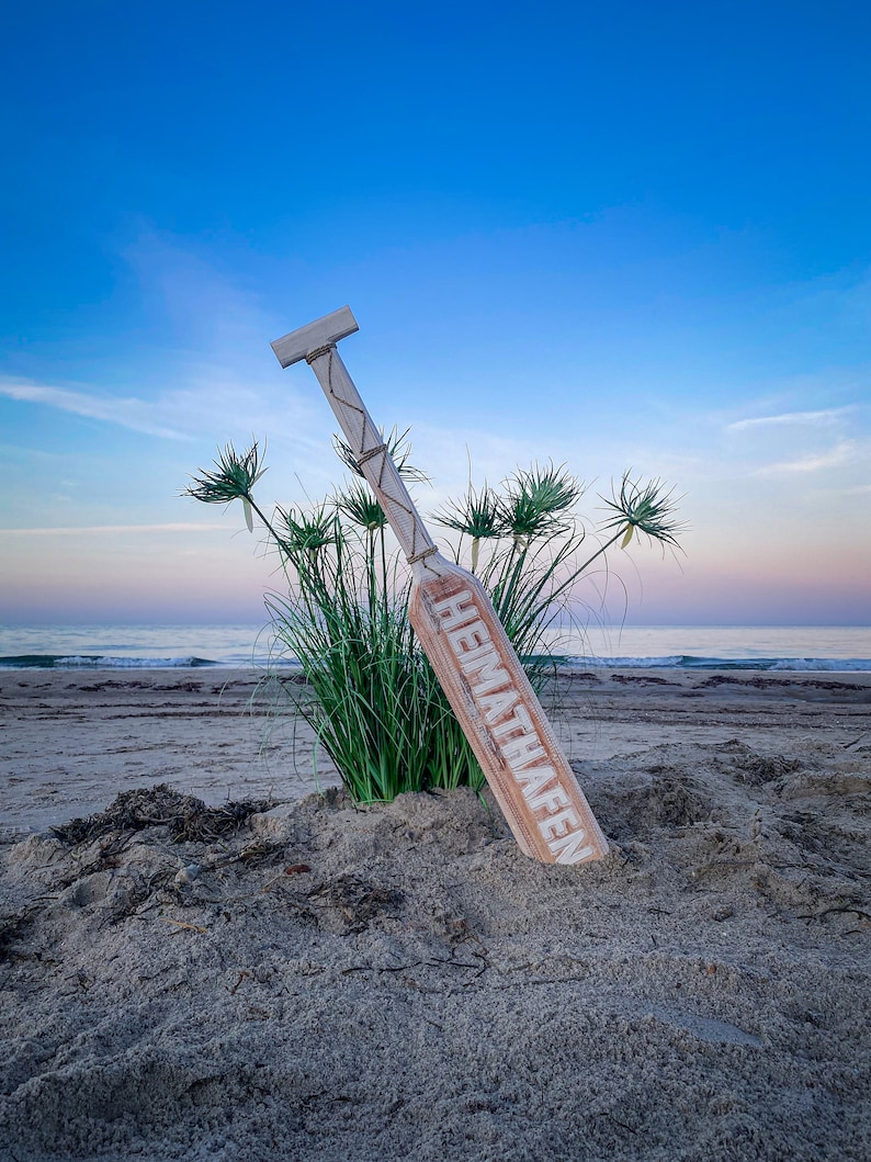 May include: A wooden sign with the word "HEIMATHAFEN" in white lettering, leaning against a patch of green grass on a sandy beach. The sky is a gradient of blue and pink, with the ocean visible in the background.