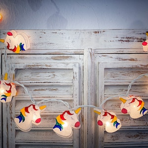 May include: A string of unicorn-shaped fairy lights. Each light features a white unicorn head with a yellow horn, pink mouth, and a rainbow mane. The lights are strung on a white wire, perfect for a child's bedroom or party decor.