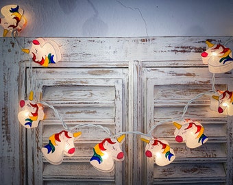 May include: A string of unicorn-shaped fairy lights. Each light features a white unicorn head with a yellow horn, pink mouth, and a rainbow mane. The lights are strung on a white wire, perfect for a child's bedroom or party decor.
