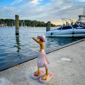 Op de afbeelding: Een houten eendenbeeldje met een roze en wit gestreepte badpak, een bijpassende hoed en slippers. De eend staat op een betonnen pier met boten en water op de achtergrond. De lucht is blauw met wolken.