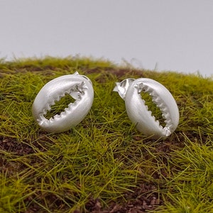 May include: A pair of silver-colored claw-shaped earrings. The earrings have a textured, matte finish and a detailed claw design. They are displayed on a bed of green and brown faux grass.