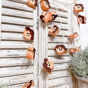May include: A string of decorative lights featuring wooden monkey faces with brown and tan details. The lights are strung on a white wire and are displayed against a white, distressed shutter background. The overall theme is whimsical and playful.