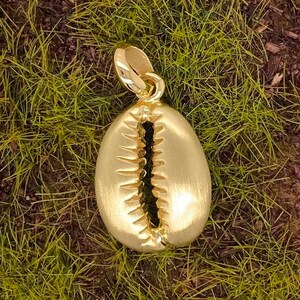 May include: A gold-colored cowrie shell pendant. The pendant has a textured surface and a detailed slit down the center. The bail is a simple loop for attaching to a chain. The pendant is set against a background of green grass.