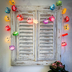 May include: A string of fairy lights with frog-shaped bulbs in various colors, including yellow, orange, red, pink, purple, blue, green, and white. The lights are strung across a white wooden window shutter.