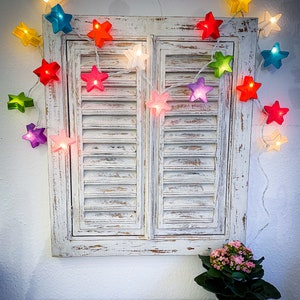 May include: A string of colorful star-shaped lights is draped over a distressed white shutter. The stars are various colors, including red, pink, green, and blue. A small potted plant with pink flowers sits below the shutter.