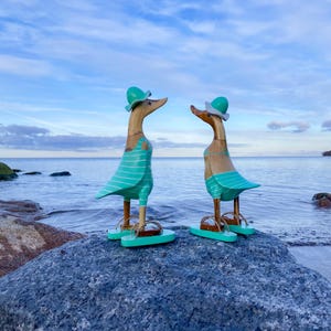 May include: Two wooden duck figurines, each wearing a teal swimsuit, hat, and sandals, stand on a rock near the water. The ducks face each other, set against a backdrop of the ocean and a cloudy sky. The ducks are approximately 25 cm tall.