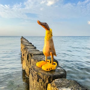 May include: A wooden duck figurine, wearing a yellow scarf and yellow clogs, stands on a wooden pier. The duck has a brown head and a light brown body. The background shows a blue sky and the sea.