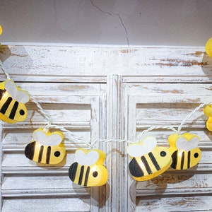 May include: A string of white lights with yellow and black bee-shaped paper lanterns. The lights are strung across a white wooden shuttered window.