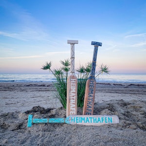 May include: Wooden decorative paddles with the text "HEIMATHAFEN" in blue and white. Two paddles stand upright, and one lies flat on the sand. The background features a beach, ocean, and a blue sky.