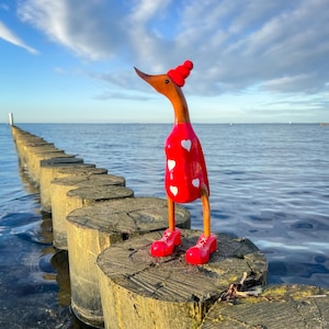 May include: A decorative wooden duck figurine, wearing a red top with white hearts, a red hat, and red shoes. The duck stands on a wooden post in the water, with the sea and sky in the background.