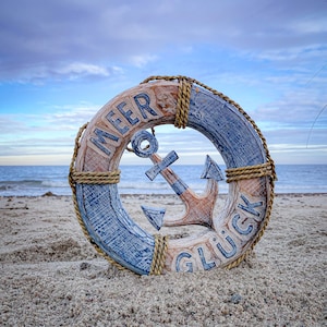 Könnte beinhalten: Eine nautische Holzdekoration in Form einer Rettungsboje mit einem Anker in der Mitte. Die Rettungsboje ist blau und beige mit den Worten "MEER GLÜCK" in Blau. Die Dekoration befindet sich an einem Sandstrand mit dem Meer im Hintergrund.