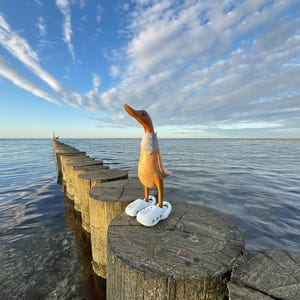 May include: A wooden duck figurine with a brown body, orange beak, and white sandals stands on a wooden post in the water. The duck wears a patterned neckerchief. The background features a blue sky with clouds and a calm body of water.