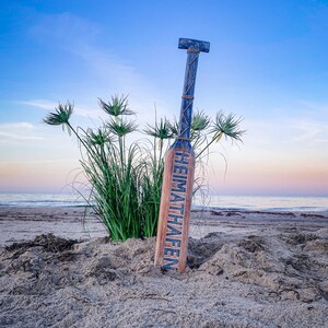 May include: A wooden paddle with the word "HEIMATHAFEN" in blue lettering, leaning in the sand on a beach. The paddle is blue and brown, with rope detailing. Green plants and the ocean are in the background under a blue sky.