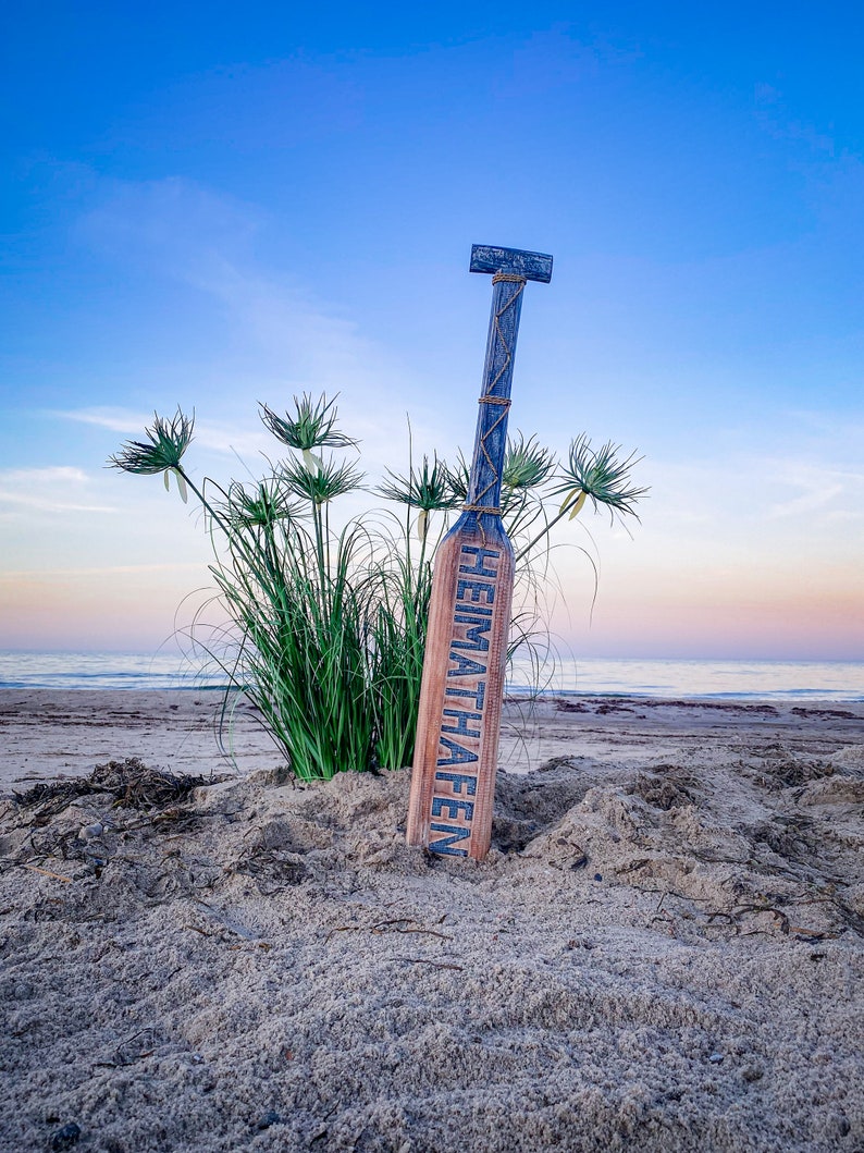 May include: A wooden paddle with the word "HEIMATHAFEN" in blue lettering, leaning in the sand on a beach. The paddle is blue and brown, with rope detailing. Green plants and the ocean are in the background under a blue sky.