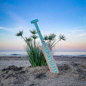 May include: A wooden paddle with a turquoise handle and accents, leaning in the sand on a beach. The paddle has the word "HEIMATHAFEN" printed on it in turquoise. Tall green grass and the ocean are in the background under a blue sky.