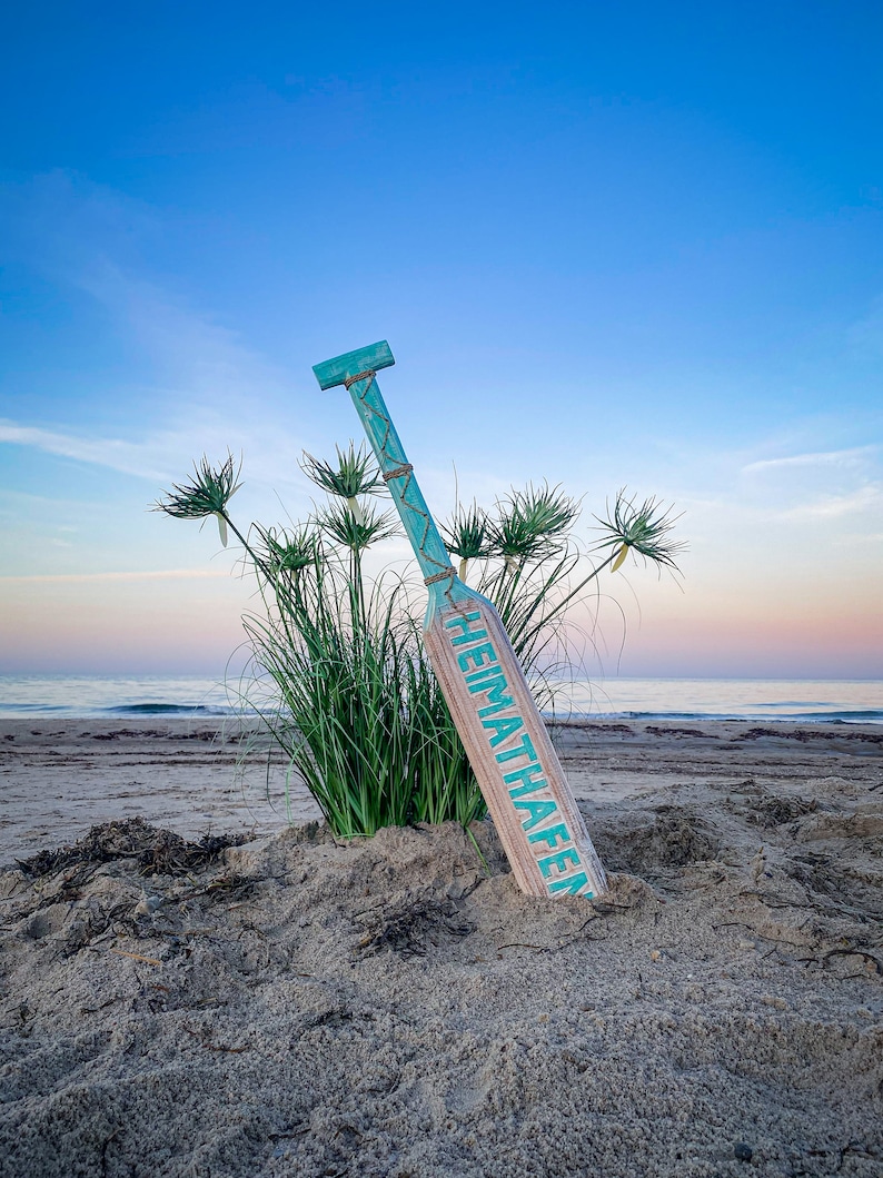 May include: A wooden paddle with a turquoise handle and accents, leaning in the sand on a beach. The paddle has the word "HEIMATHAFEN" printed on it in turquoise. Tall green grass and the ocean are in the background under a blue sky.