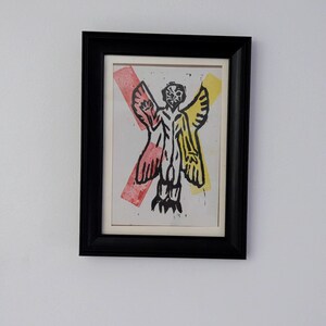 May include: A framed print depicting a stylized figure with wings, in black, yellow, and red. The artwork is set against a white background, framed in a black rectangular frame, and mounted on a white wall.