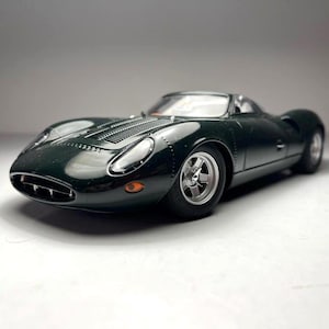 May include: A dark green vintage model sports car with silver wheels. The car has a sleek, aerodynamic design with a low profile and a curved windshield. The model car is a detailed replica of a classic vehicle.