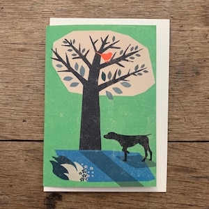 May include: A green and cream card featuring a black silhouette of a tree with a red bird perched on a branch. A black dog is standing in the grass below the tree.