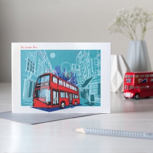Puede incluir: Una tarjeta de felicitaci&oacute;n con una ilustraci&oacute;n de un autob&uacute;s londinense de dos pisos rojo sobre un fondo turquesa con dibujos de edificios en blanco. El texto "The London Bus" est&aacute; en la parte superior izquierda. Un peque&ntilde;o modelo de autob&uacute;s rojo est&aacute; en el fondo.