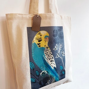 May include: A white canvas tote bag featuring a blue and yellow parakeet illustration. The parakeet is perched on a branch with blue and green leaves. The bag has brown straps.